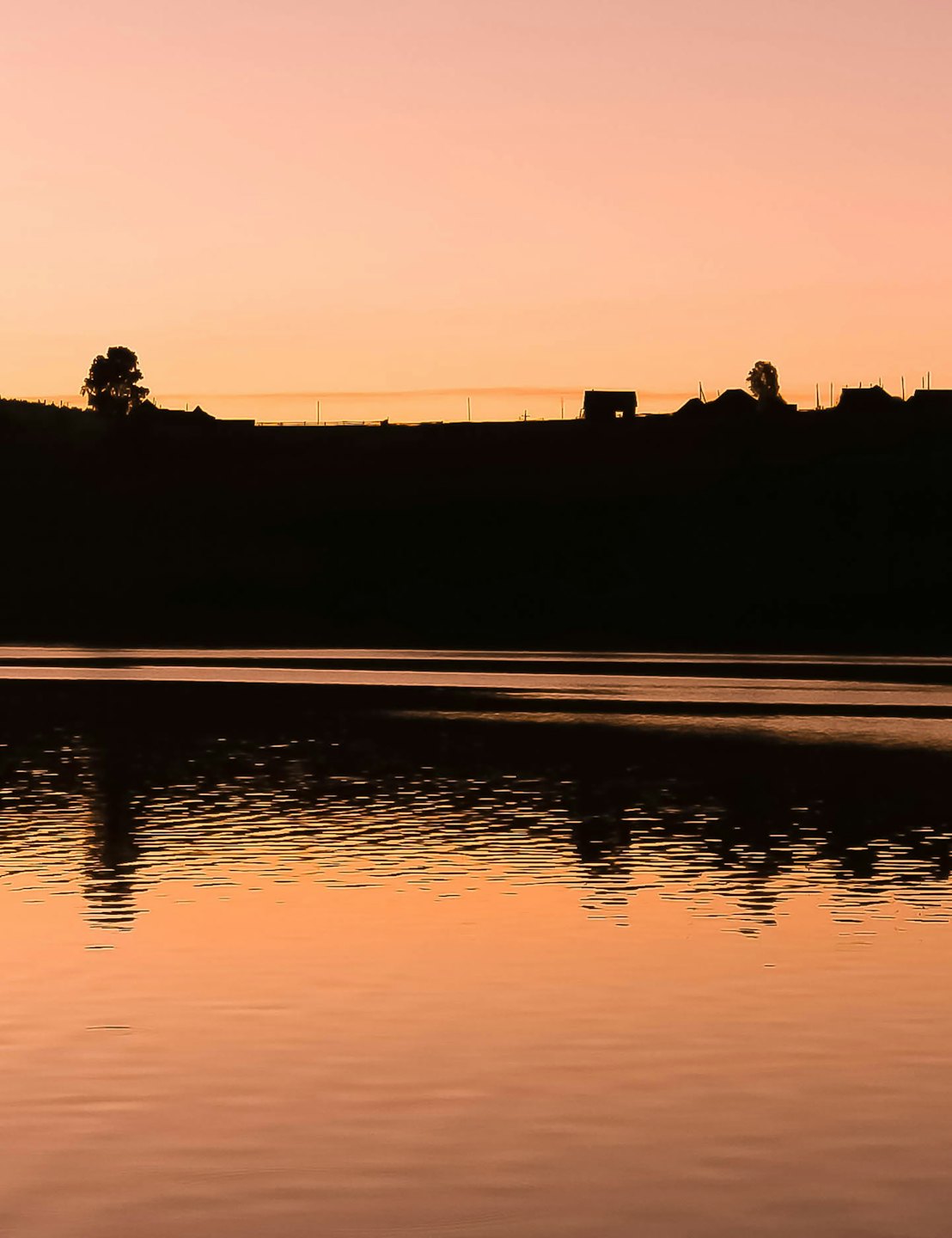 Distant hill and structures reflected in calm water at twilight.